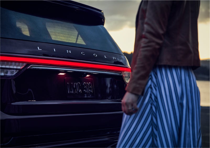 A person is shown near the rear of a 2023 Lincoln Aviator® SUV as the Lincoln Embrace illuminates the rear lights | Haldeman Lincoln in Allentown PA